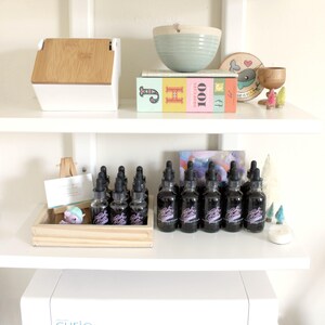 May include: A white shelf with a variety of items on it, including a wooden box, a book titled "100 Beautiful Stitches", a bowl, a wooden sign with a narwhal, and a collection of small glass bottles with droppers. The bottles are arranged in a row on the shelf and are filled with a dark liquid. The bottles have a purple label with the text "Baby Got Booze" on them.