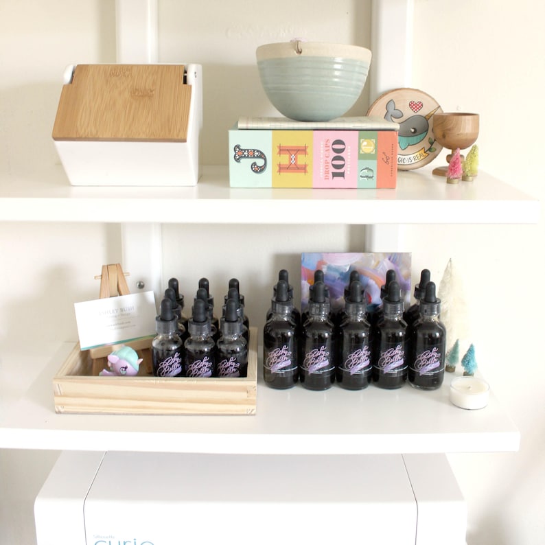 May include: A white shelf with a variety of items on it, including a wooden box, a book titled "100 Beautiful Stitches", a bowl, a wooden sign with a narwhal, and a collection of small glass bottles with droppers. The bottles are arranged in a row on the shelf and are filled with a dark liquid. The bottles have a purple label with the text "Baby Got Booze" on them.