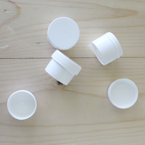 Peut inclure: Cinq contenants en plastique blanc avec des couvercles. Les contenants sont de différentes tailles et formes. Ils sont sur une surface en bois clair.