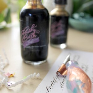 May include: Two glass bottles of black ink with a gold and black calligraphy pen. The bottles are labeled "Fussy Broth" in pink lettering. The pen is resting on a piece of paper with calligraphy writing.