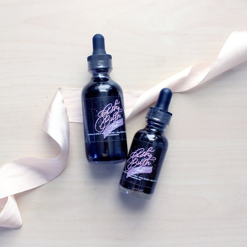 May include: Two small glass bottles of black ink with a white label that says "Echo Butter Ink". The bottles have a dropper top and are sitting on a light wood surface with a light pink ribbon.