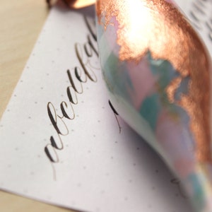 May include: Close-up of a copper-toned glass ornament with a swirling, marbled pattern in shades of pink, blue, and white. The ornament is lying on a sheet of paper with a handwritten inscription in black ink.