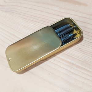 May include: A gold metal tin with a hinged lid, open to reveal a set of five black calligraphy nibs.