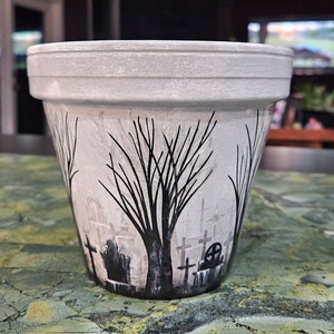 Può includere: Un vaso di ceramica bianco con un disegno dipinto in bianco e nero di un cimitero con alberi. Il vaso è appoggiato su una superficie verde e marrone.