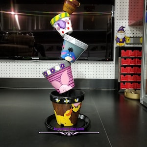 Pode incluir: Uma pilha de cinco vasos de flores de terracota pintados à mão e decorados com personagens do Winnie the Pooh. Os vasos estão empilhados em forma de torre, com o vaso superior apresentando um pote de mel com as palavras "Hunny". O vaso inferior apresenta o Winnie the Pooh, o Tigrão e o Porquinho. Os vasos são uma forma divertida e caprichosa de adicionar um toque de magia Disney à sua casa.
