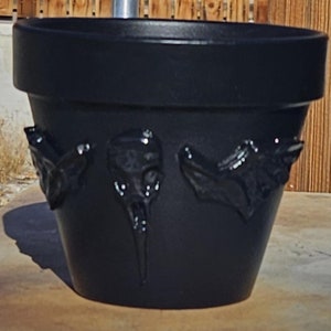 以下が含まれることがあります： 3つの浮き彫りの彫刻が施された黒いテラコッタ製のプランター。プランターは木製の表面の上に置かれています。