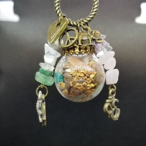Pode incluir: Um colar de ouro com um frasco de vidro cheio de pequenas pedras e cristais. O frasco é rodeado por pingentes, incluindo um sinal de paz, um elefante e um símbolo Om.