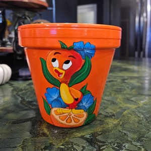 Pode incluir: Pote de flores de terracota laranja com um desenho pintado à mão de um pássaro laranja de desenho animado com flores azuis e uma fatia de laranja.