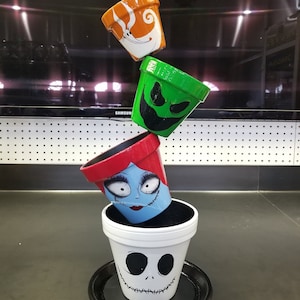 Pode incluir: Uma pilha de cinco vasos de flores de terracota pintados com desenhos de personagens de Halloween. O vaso do topo é roxo com um personagem preto e branco, o segundo vaso é laranja com um personagem branco, o terceiro vaso é verde com um personagem preto, o quarto vaso é vermelho e azul com um personagem branco, e o vaso do fundo é branco com um personagem preto.