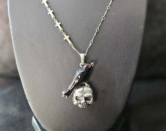 May include: A silver chain necklace with a pendant featuring a black crow perched on a silver skull. The crow has red eyes.