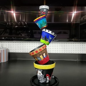 Pode incluir: Uma pilha de cinco vasos de flores pintados com diferentes desenhos. O vaso superior é vermelho com estrelas brancas e o texto "There's No Place Like Home". O segundo vaso é azul com um desenho branco. O terceiro vaso é verde com um desenho branco. O quarto vaso é vermelho com um desenho branco. O vaso inferior é preto com uma faixa amarela e um desenho branco.