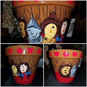 May include: A brown terracotta flower pot with a wood grain design painted on the side. The pot is decorated with red apples around the top rim and a painted scene of the characters from the Wizard of Oz. The characters include Dorothy, the Scarecrow, the Tin Man, and the Cowardly Lion.