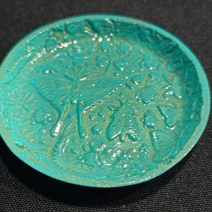 May include: A round, turquoise-colored dish with an embossed design featuring a butterfly, insects, and floral elements. The dish has a slightly aged, metallic finish, and is set against a dark background. The decorative dish is likely for holding small items.