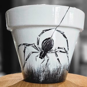 May include: White ceramic plant pot featuring a hand-painted black spider. The spider design is the focal point, with its legs and web painted across the pot's surface. The base of the pot has a textured, dark grey and white painted effect.