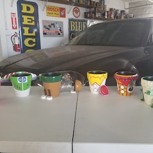 May include: Six painted flower pots, each decorated with a different character from the Toy Story movie franchise. The pots are arranged in a row on a white surface. The characters include Woody, Buzz Lightyear, Rex, Hamm, Slinky Dog, and Mr. Potato Head.