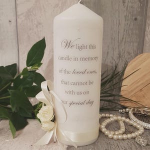 May include: A white pillar candle inscribed with the words "We light this candle in memory of the loved ones, that cannot be with us on our special day." It is decorated with a white ribbon and roses. The background includes greenery and jewellery.