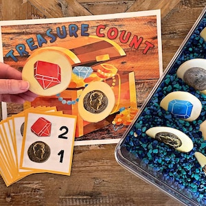 May include: A colorful learning game for kids featuring a treasure chest with gems and coins. The game includes a set of cards with numbers and images of red gems and gold coins. The game is designed to help children learn to count.