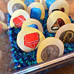 May include: A clear plastic container filled with blue and green pebbles.  The container holds several round, yellow tokens with various images on them, including red and blue gems, rings, and coins.