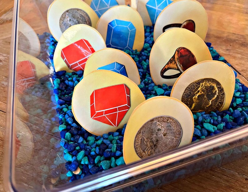 May include: A clear plastic container filled with blue and green pebbles.  The container holds several round, yellow tokens with various images on them, including red and blue gems, rings, and coins.
