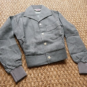 May include: A gray and white checkered jacket with a button-up front and long sleeves. The jacket has a small pocket on the left side and a label that says "Wilton" on the inside collar.