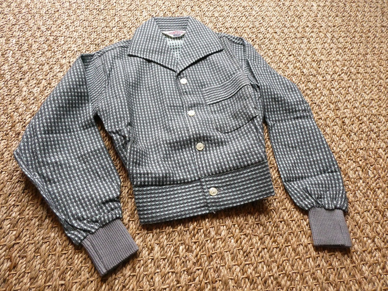 May include: A gray and white checkered jacket with a button-up front and long sleeves. The jacket has a small pocket on the left side and a label that says "Wilton" on the inside collar.