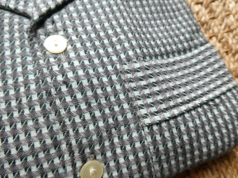 May include: A close-up of a black and light blue patterned shirt with a pocket on the left side. The shirt has a button closure and a collar.