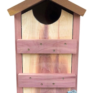 May include: A wooden birdhouse with a cedar wood exterior and a brown cedar wood roof. The birdhouse has a hinged door with a latch and two wooden shelves inside.