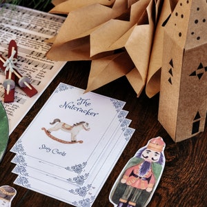 Puede incluir: Un conjunto de tarjetas de cuentos de "El Cascanueces" con una ilustración de un caballo balancín y bordes azules decorativos. También se ve un recorte de papel de un soldado Cascanueces y una casa de papel marrón, junto con partituras.