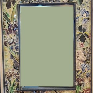 May include: A decorative mirror with a mosaic frame featuring a floral design. The frame is made of small pieces of glass and ceramic in shades of green, blue, purple, and white. The mirror is surrounded by a black frame.