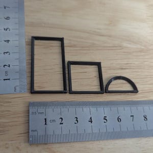 May include: Three small, dark brown geometric shapes: a rectangle, a square, and a semi-circle. A ruler with centimetre and millimetre markings is placed next to the shapes for scale. The shapes appear to be 3D printed.