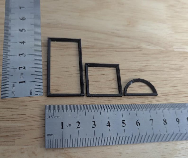 May include: Three small, dark brown geometric shapes: a rectangle, a square, and a semi-circle. A ruler with centimetre and millimetre markings is placed next to the shapes for scale. The shapes appear to be 3D printed.