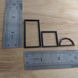 May include: Four small, black, geometric shapes: a rectangle, a square, and a half-circle, are arranged on a wooden surface. A metal ruler with inch markings is positioned next to the shapes for scale. The shapes appear to be 3D printed.