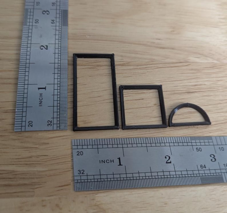 May include: Four small, black, geometric shapes: a rectangle, a square, and a half-circle, are arranged on a wooden surface. A metal ruler with inch markings is positioned next to the shapes for scale. The shapes appear to be 3D printed.