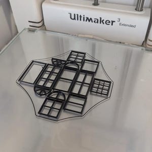 May include: A black plastic 3D-printed design featuring various window shapes and styles. The design includes rectangular, arched, and grid-like window frames. The object is on a clear surface, with an Ultimaker 3 Extended 3D printer in the background.