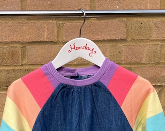 May include: A denim dress with rainbow-striped sleeves hangs on a white hanger. The sleeves feature horizontal stripes of pink, orange, yellow, light blue, and purple. The hanger has the word "Monday" written on it in red.