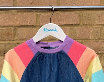 May include: A long-sleeved dress featuring a dark blue denim bodice and a drop-waist skirt. The sleeves are designed with horizontal stripes in shades of pink, orange, yellow, light blue, and purple. The dress hangs on a white hanger labeled Hannah.