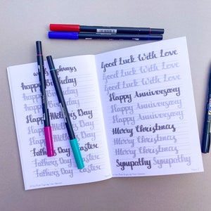 May include: An open notebook with lined pages filled with handwritten calligraphy practice. The notebook is open to a page with the words "Good Luck with Love", "Happy Anniversary", "Merry Christmas", and "Sympathy" written in different styles. There are several black, blue, and pink markers on the table next to the notebook.