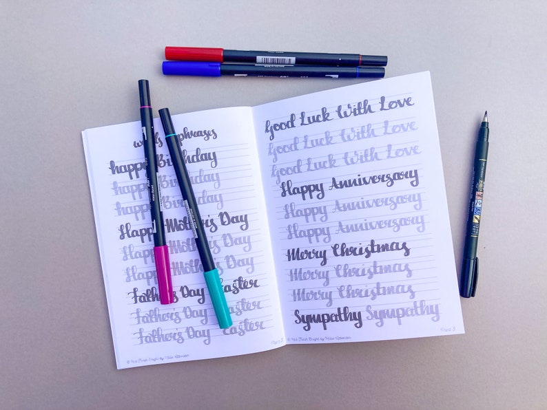 May include: An open notebook with lined pages filled with handwritten calligraphy practice. The notebook is open to a page with the words "Good Luck with Love", "Happy Anniversary", "Merry Christmas", and "Sympathy" written in different styles. There are several black, blue, and pink markers on the table next to the notebook.