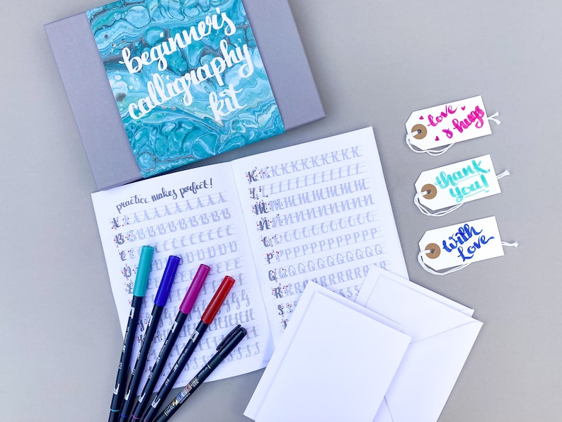 May include: A beginner's calligraphy kit with a teal and blue marbled cover, a practice booklet, five calligraphy pens, three blank cards, and three gift tags with the words "love & hugs", "thank you", and "with love" written in calligraphy.