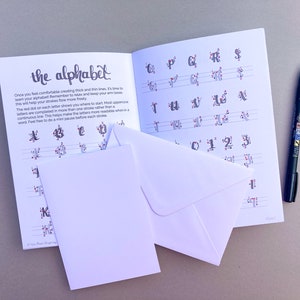 May include: A workbook with the title "the alphabet" on the cover. The book has red dots on the pages to help guide the user in creating letters. The book is open to a page with instructions on how to create letters. There are two white envelopes on top of the book. A black pen is lying on the table next to the book.