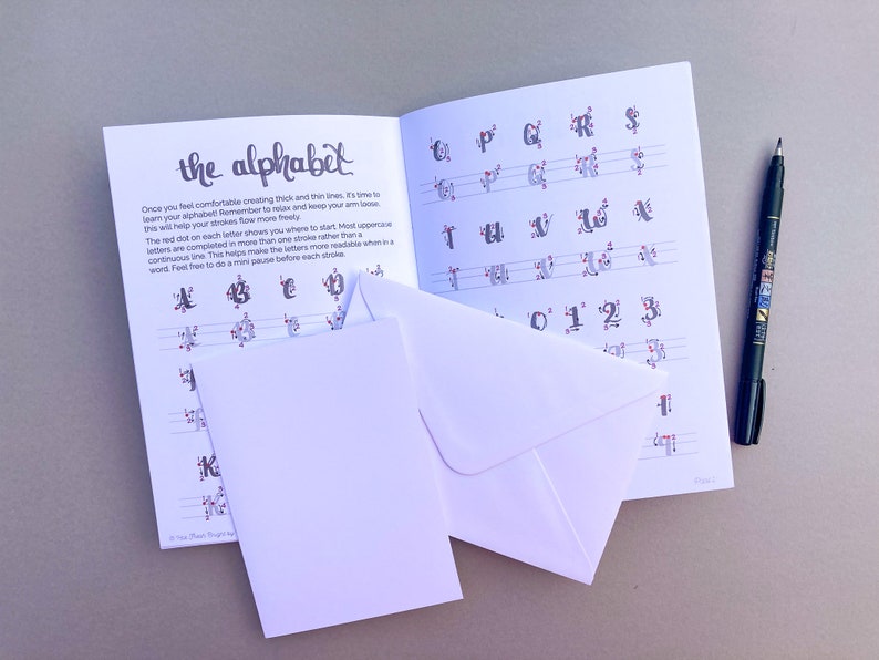 May include: A workbook with the title "the alphabet" on the cover. The book has red dots on the pages to help guide the user in creating letters. The book is open to a page with instructions on how to create letters. There are two white envelopes on top of the book. A black pen is lying on the table next to the book.