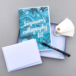 May include: A calligraphy kit with a blue and green marbled design booklet, a black pen, and a stack of white paper cards. The booklet has the text "beginner's calligraphy kit" on the front.