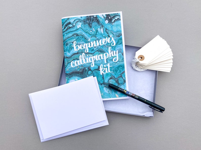 May include: A calligraphy kit with a blue and green marbled design booklet, a black pen, and a stack of white paper cards. The booklet has the text "beginner's calligraphy kit" on the front.