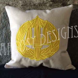 May include: A white pillow with a yellow embroidered design of a trident and laurel wreath. The design is the official emblem of the United States Navy SEALs.