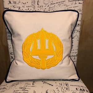 May include: A white pillow with a navy blue border features an embroidered yellow trident and laurel wreath.