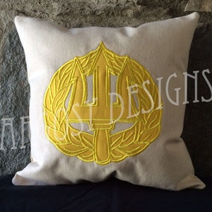 May include: A white pillow with a yellow embroidered trident and laurel wreath. The trident is a symbol of the United States Navy SEALs.