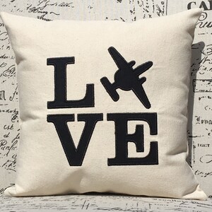 May include: A white throw pillow with black embroidered text that reads "LOVE" with a small airplane graphic between the "L" and the "O".