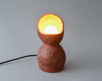 May include: A terracotta-colored table lamp with a textured surface. The lamp has a round, bulbous base and a wide, open top. A light bulb is visible inside the lamp, illuminating the interior.