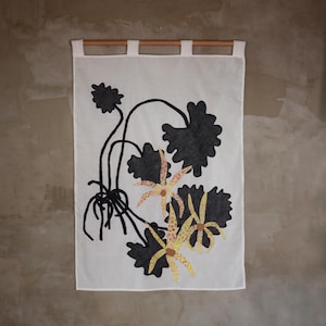 May include: A white rectangular fabric wall hanging suspended from a wooden rod. The textile features a screen-printed botanical illustration of dark black scalloped leaves and three yellow flowers with brown spots and thin, elongated petals.