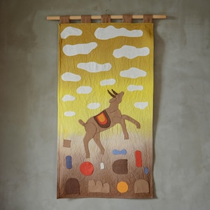 May include: A fabric wall hanging with a brown goat leaping against a yellow and brown gradient background with white cloud shapes. The goat has a saddle and is set against a sky with white clouds. The bottom has abstract shapes.